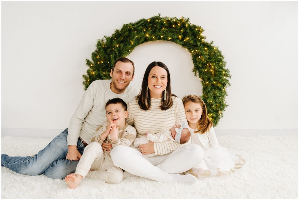 smiling family photo in front of christmas wreath with newborn baby